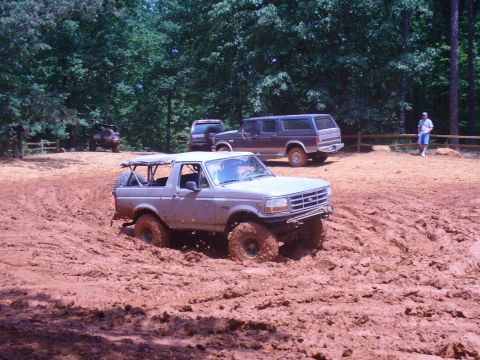 1996 Ford Bronco - A silver convertible SUV navigates a muddy off-road course, with other vehicles and a person visible in the background.