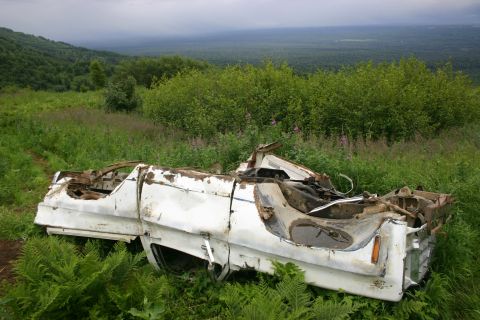 2003 Jeep TJ - A severely damaged white pickup truck sits abandoned in a grassy field, with a dense forest and rolling hills visible in the distance.