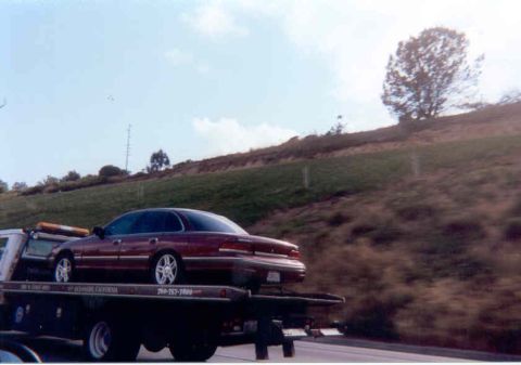 1992 Ford Crown Victoria - Dark burgundy Ford Crown Victoria sedan strapped to a black flatbed tow truck, shot from the passenger rear three-quarter as the rig rolls…