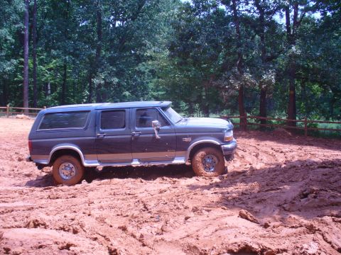 1996 Ford Bronco - A Ford Bronco navigates a muddy off-road course, with trees and a wooden fence visible in the background.