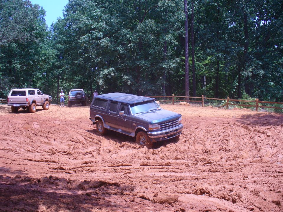 Dark blue Ford Bronco stuck in mud, front 3/4 view. Muddy terrain and trees in background