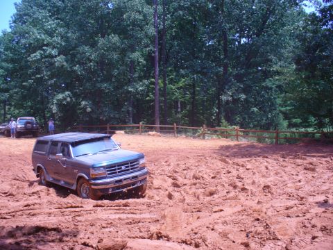 1996 Ford Bronco - A Ford Bronco navigates a muddy off-road course with trees and a wooden fence visible.
