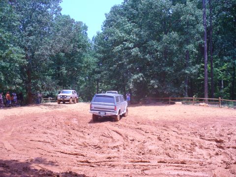 1996 Ford Bronco - Two off-road vehicles navigate a muddy track surrounded by trees.