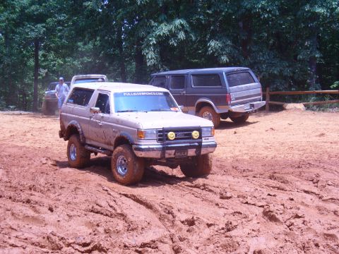 1996 Ford Bronco - Two Ford Broncos navigate a muddy off-road trail surrounded by trees.
