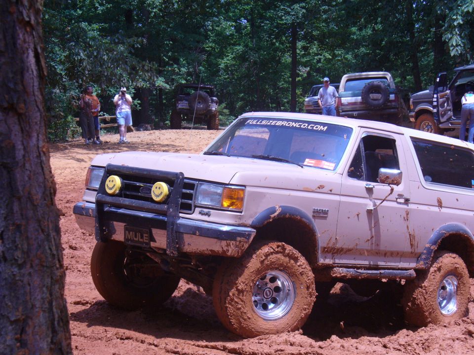 Tan Ford Bronco with smiley face lights on bull bar stuck in mud, other vehicles and people in background