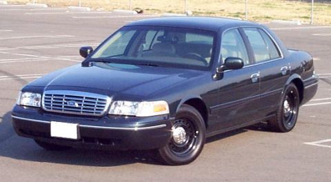 1998 Ford Crown Victoria - A dark-colored sedan is parked in an asphalt lot under a bright sky.