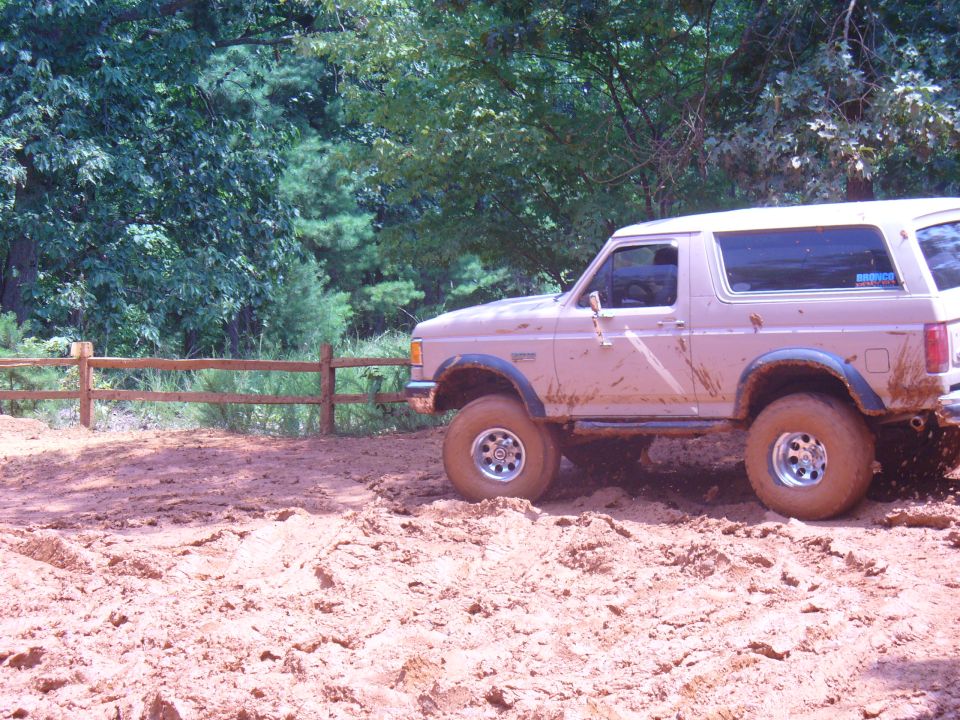 Tan Ford Bronco covered in mud, driving through a dirt track with trees in the background