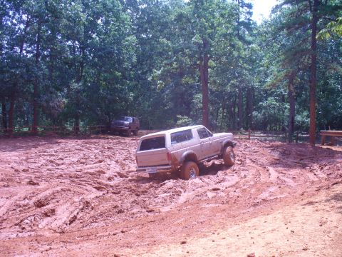 1996 Ford Bronco - Two SUVs navigate a muddy off-road course with trees surrounding the area.
