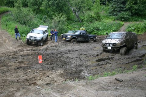 2003 Jeep TJ - Multiple off-road vehicles are stuck in deep mud on a trail surrounded by trees.