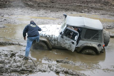 2003 Jeep TJ - A white 2003 Jeep TJ is stuck deep in mud, with a person attempting to help push it free.