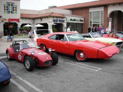 1998 Ford Crown Victoria - red Lotus-style racing roadster numbered 29 sits beside a red Dodge Charger Daytona outside a strip-mall coffee shop.