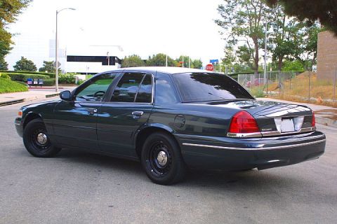 1998 Ford Crown Victoria - A dark sedan is viewed from the rear quarter, parked on a paved surface with trees and buildings in the background.