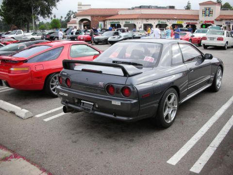 1998 Ford Crown Victoria - gray Nissan Skyline GT-R seen from the rear with its tall wing and round tail lights, parked among other cars at a show.
