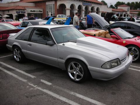 1998 Ford Crown Victoria - silver Fox-body Ford Mustang 5.0 hatchback on polished wheels, parked among other cars at a strip-mall show.