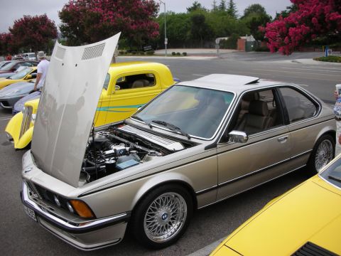 1998 Ford Crown Victoria - gold BMW 6-Series coupe with its hood raised showing the engine, parked next to a yellow hot rod at a car show.