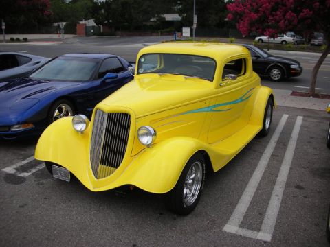 1998 Ford Crown Victoria - bright-yellow 1930s Ford three-window hot rod coupe with a blue pinstripe and chrome wheels, parked beside a blue Corvette.