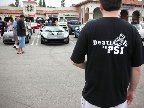 1998 Ford Crown Victoria - man wearing a black 'Death by PSI' T-shirt stands facing a black Ford GT and crowd at a strip-mall car show.