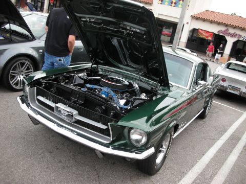 1998 Ford Crown Victoria - dark green vintage Ford Mustang fastback sits with its hood raised, exposing a chrome-dressed V8 engine, at a sunny lot car show.