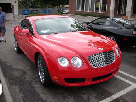 1998 Ford Crown Victoria - bright red Bentley Continental GT coupe with its mesh grille and round quad headlights sits in a parking lot beside a dark sports car.