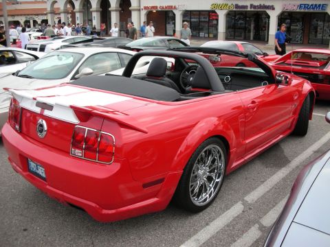 1998 Ford Crown Victoria - red Ford Mustang convertible with a body kit, white racing stripes, and chrome wheels sits top-down in a busy car-show lot.