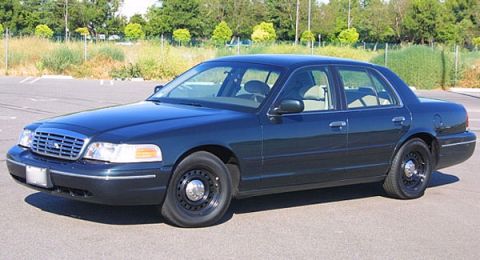 1998 Ford Crown Victoria - dark green Ford Crown Victoria sedan with black steel wheels parked, viewed from the front left three-quarter near a fence.