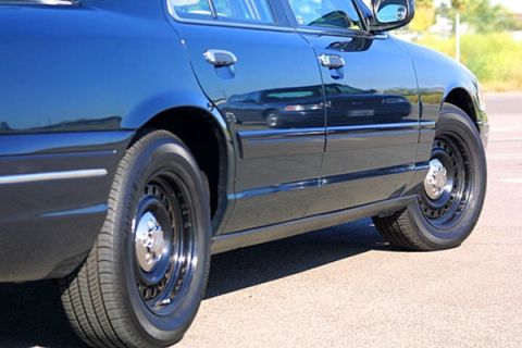 1998 Ford Crown Victoria - Low close-up of a dark sedan's lower body panels and rear steel wheel with chrome trim ring, taken in bright sun.