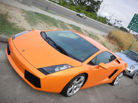 1998 Ford Crown Victoria - orange Lamborghini Gallardo sits at the roadside with chrome wheels, a silver Porsche visible alongside it.