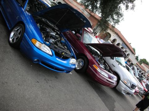 1998 Ford Crown Victoria - blue Ford Mustang and a maroon sedan stand with hoods raised among a row of show cars near a tile-roofed plaza.