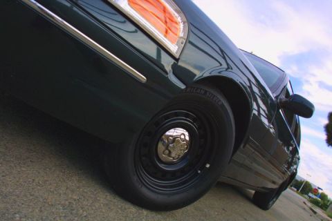 1998 Ford Crown Victoria - Dramatic low-angle close-up of a dark sedan's rear quarter showing the steel wheel, tire sidewall and red taillight against sky.