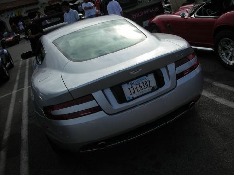 1998 Ford Crown Victoria - rear of a silver Aston Martin coupe shows its taillights and a Montana license plate in a busy show lot.