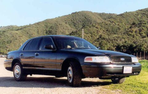 1998 Ford Crown Victoria - dark Ford Crown Victoria sedan parked on gravel in front of brush-covered hills, viewed from the front right three-quarter.