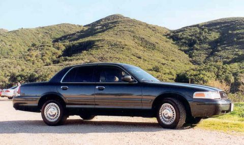 1998 Ford Crown Victoria - Side profile of a dark Ford Crown Victoria sedan parked on gravel with brush-covered hills rising behind it.