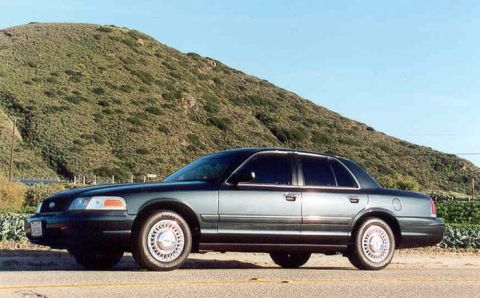 1998 Ford Crown Victoria - Side profile of a dark green Ford Crown Victoria sedan with alloy-look wheels parked along a road with a grassy hill behind.