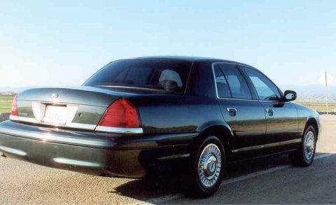 1998 Ford Crown Victoria - dark Ford Crown Victoria sedan with chrome wheels viewed from the rear left three-quarter in a flat open landscape under blue sky.