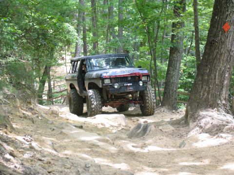 1995 Ford C-350 (Centurion Conversions) - A heavily modified SUV navigates a challenging off-road trail surrounded by dense woods.