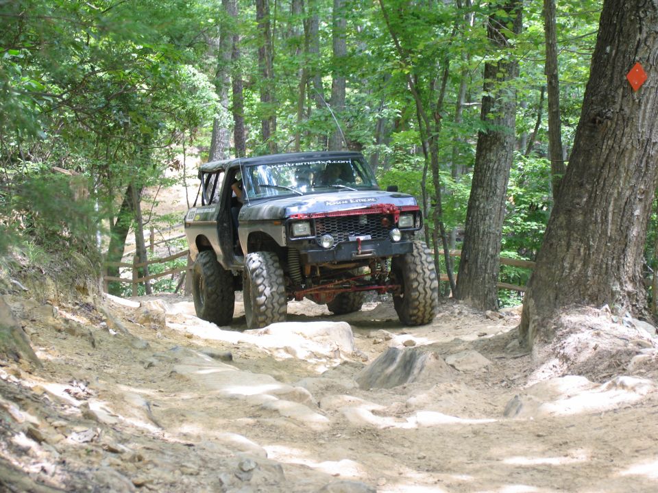 Modified off-road SUV with large tires driving on a dirt and rock trail through a forest