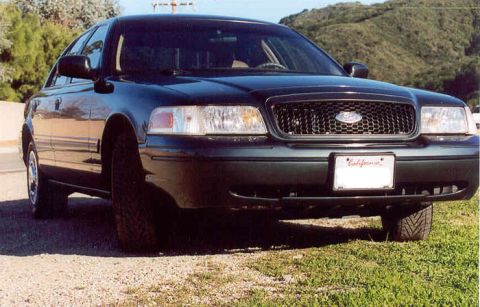 1998 Ford Crown Victoria - Front view of a dark Ford Crown Victoria sedan showing its grille and headlights, with a California license plate, hills behind.