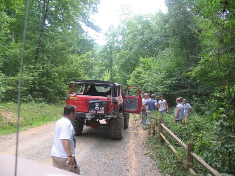 1995 Ford C-350 (Centurion Conversions) - A group of people gather around a modified red off-road vehicle on a wooded dirt path.