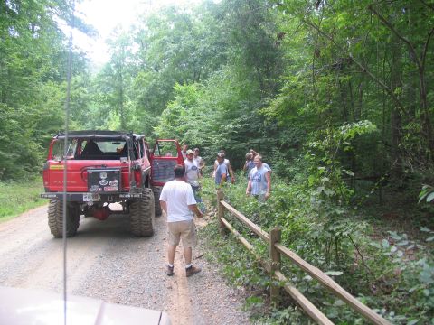 1995 Ford C-350 (Centurion Conversions) - A group of people stand around a modified red Ford Bronco on a dirt road in a wooded area.