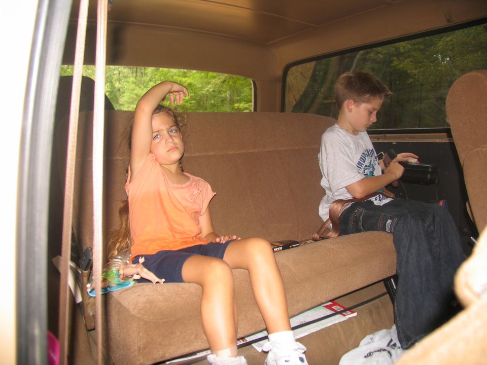 Two children sitting in the back seat of a vehicle, one looking bored, the other playing with a device