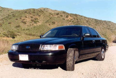 1998 Ford Crown Victoria - black Ford Crown Victoria sedan viewed from the front left three-quarter, parked on gravel before brush-covered hills.