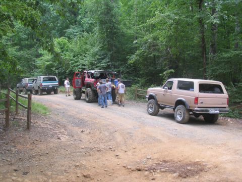 1995 Ford C-350 (Centurion Conversions) - A group of off-road vehicles and people are gathered on a dirt road in a wooded area.