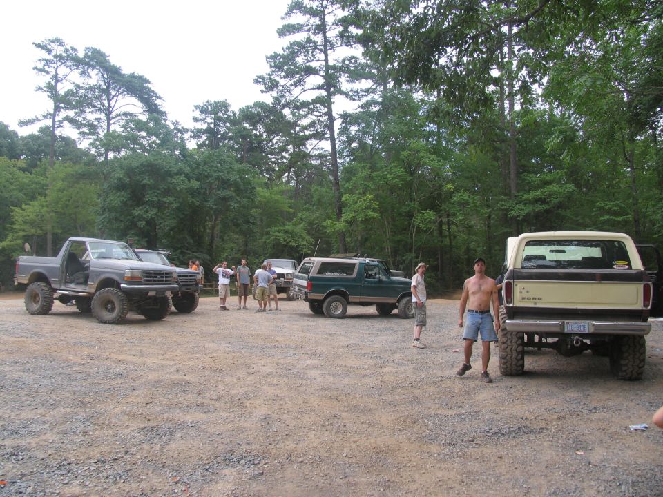 Several off-road trucks and SUVs parked in a gravel lot with trees in the background. People are standing around the vehicles