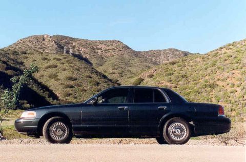 1998 Ford Crown Victoria - Side profile of a dark Ford Crown Victoria sedan with chrome wheels parked on a dirt shoulder with green hills rising behind.