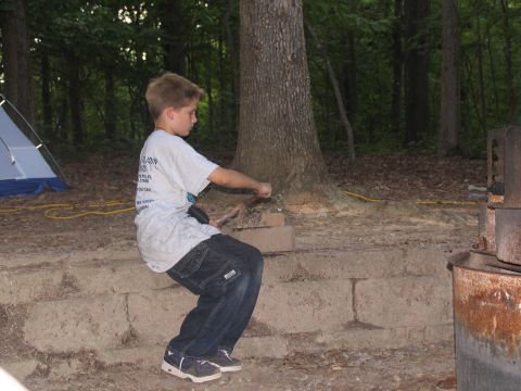 1995 Ford C-350 (Centurion Conversions) - A young boy sits on a stone wall in a wooded area, with a tent and campfire pit visible.