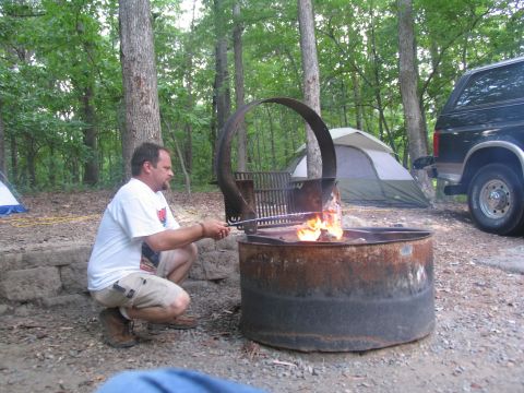 1995 Ford C-350 (Centurion Conversions) - A man cooks over a campfire at a campsite, with tents and a vehicle nearby.