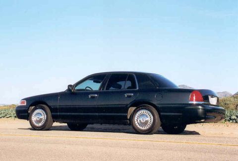1998 Ford Crown Victoria - black Ford Crown Victoria sedan with chrome wheels viewed from the rear left three-quarter on a road in a flat open landscape.