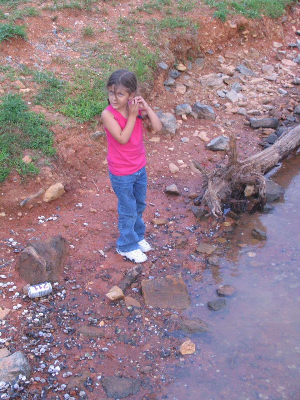 Girl in pink shirt and jeans by a muddy bank with rocks and a log