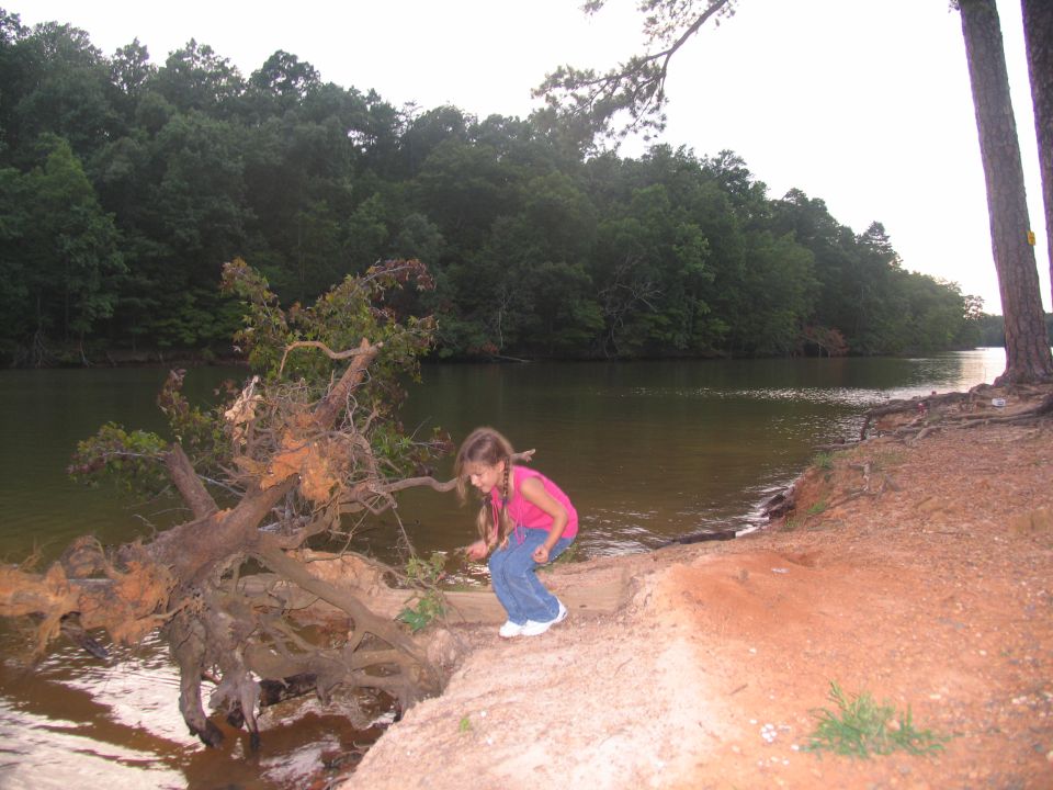 Girl in pink shirt and jeans crouches on a sandy bank next to a lake with a fallen tree