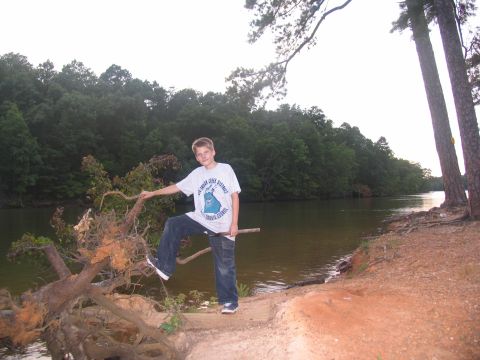 1995 Ford C-350 (Centurion Conversions) - A young boy poses on a fallen tree near the water's edge, with a dense forest lining the opposite bank.